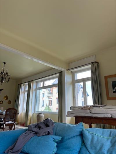 The image shows a living room in an apartment in Dinard, France, located at coordinates 48.638, -2.057. The room has pale yellow walls, a white ceiling, and large windows with white curtains. There is a blue couch in the foreground with a gray sweater on top of it. The windows look out onto an urban scene with a building in the background. A wooden table is in the right of the image, with a stack of white cushions on top of it. A chandelier hangs from the ceiling in the background. The scene is mostly empty, suggesting that no one is in the room at this time.