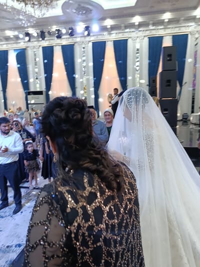 A wedding is taking place in a large, ornate hall in Shahrisabz, Uzbekistan. In the foreground, a woman with dark, curly hair is seen from behind, wearing a black dress adorned with gold sequins. She is facing a bride, who is wearing a white wedding gown and a long, sparkling veil. The bride is also facing away from the camera, with her head bowed. In the background, a crowd of guests are gathered, some dressed in traditional attire. There are also large speakers and stage lighting visible, suggesting a performance or ceremony is underway. The hall itself appears to be decorated with blue drapes and white columns, creating a festive atmosphere. The floor is covered in a patterned carpet.