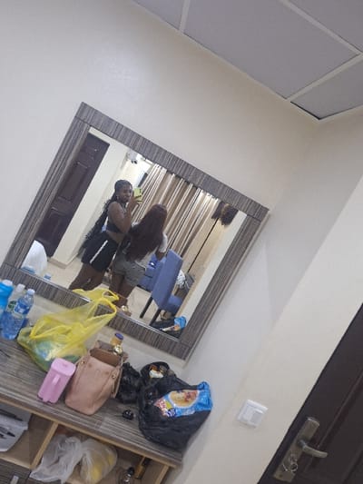 The image depicts an indoor scene within a room in Close, Nigeria. A large rectangular mirror with a wide, horizontally striped frame dominates the central upper portion of the view. Reflected in the mirror are two individuals. The person on the left, identified as a young Black female, has long dark braids, wears a dark crop top and dark shorts with a white stripe, and is holding a smartphone with a light green case to capture a mirror selfie. The second person, to her right, also a young Black female, is seen from behind, wearing a light-colored t-shirt and light-colored shorts, with long, straight dark brown hair.
Reflected in the background of the mirror are light brown vertical blinds, a dark brown wooden door with a metallic handle, and a blue upholstered chair featuring a patterned backrest. Below the mirror, a striped wooden dresser is cluttered with numerous items. These include two clear plastic bottles of water (one with a blue cap), a yellow translucent plastic bag, a pink plastic container with a handle, a light brown leather-like handbag containing a small bottle, and several black plastic bags, one of which visibly contains a blue-wrapped snack item. Sunglasses and a small black object are also present on the dresser. Additional items, including a white plastic container and more plastic bags, are visible on the dresser's lower shelves. The room features plain light-colored walls, a ceiling with rectangular paneling, and a white light switch visible on the wall to the right of the dresser. A dark brown door with a metallic handle is also partially visible on the far right side of the image, outside the mirror's reflection.