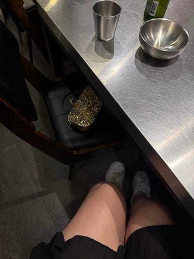 This media file captures a close-up, overhead view of a stainless steel tabletop. On the table, a metallic bowl and a smaller metallic cup are visible, along with part of a green bottle with a label. Below the table, two people's legs and feet are visible, wearing white sneakers and black clothing. A sequined handbag is also placed on the floor under the table. The scene appears to be indoors, possibly in a restaurant or bar, given the table setting and the presence of drinks. The lighting suggests it could be evening or a dimly lit indoor space. The overall mood is casual and relaxed. No specific weather conditions or explicit emotions are conveyed. No discernible text is clearly legible in the image. The location is identified as Cebu City, Philippines.