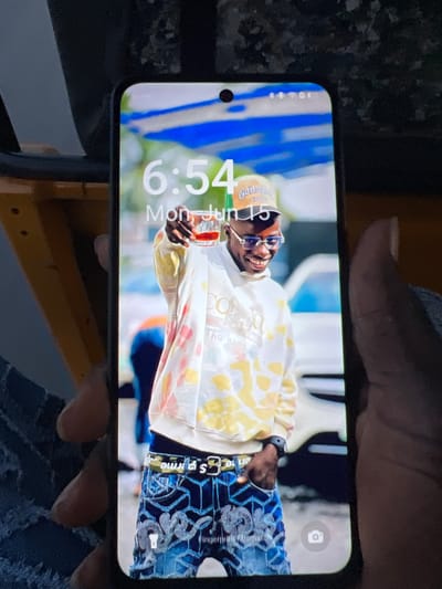 The image shows a smartphone held in a person's hand, displaying a lock screen. The lock screen features a picture of a young Black man smiling and holding up a glass of amber liquid. He is wearing a yellow and white tie-dye hoodie, blue-tinted glasses, a brown baseball cap, and detailed blue jeans. The background of the image on the phone appears to be an outdoor setting with a blue and white structure overhead.
The phone's time is displayed as 6:54, and the date is Monday, June 15. The phone also shows signal strength, battery level, and possibly other status icons in the top right corner. A camera icon is visible in the lower right corner of the phone's screen. The phone itself has a black bezel and a visible notch at the top center of the screen.
The surrounding environment of the photograph shows parts of what appears to be a yellow vehicle, possibly a tricycle or a similar mode of transport, and a dark, textured surface above the phone. The overall lighting suggests it is daytime. The image does not provide any specific visual cues to confirm the location as Orifite, Nigeria.