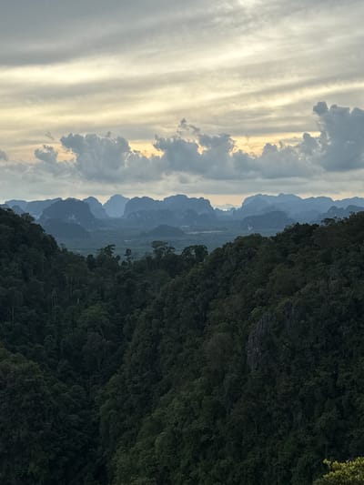 This is a vertical, landscape photograph taken from a high vantage point in Ban Khao Tang, Thailand. The image depicts a dramatic natural scene characterized by a striking contrast between a vast, cloudy sky and a lush, mountainous terrain.
On the left side of the frame, a turbulent sky dominates, filled with streaks of clouds in shades of gray, white, and soft yellow. The light suggests either sunrise or sunset, casting a warm, diffused glow through the atmospheric layers.
The right side of the image is consumed by a dense, dark green forest that clings to steep, rocky slopes. The foliage appears thick and untamed, with hints of rocky outcrops visible through the dense vegetation. This forms a natural, imposing boundary for the scene.
In the mid-ground, a series of majestic, mist-shrouded mountains recede into the distance, their peaks softened by atmospheric perspective and rendered in varying shades of blue and gray. This layered effect creates a sense of depth and vastness. Below the mountains, a valley floor is visible, hinting at human settlement or agricultural land, though it is not clearly defined.
The overall mood of the image is serene yet powerful, capturing the grandeur of the natural landscape at a transitional time of day. There are no people, objects, or visible text in the photograph. The weather appears to be partly cloudy, contributing to the dramatic sky.