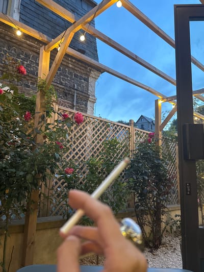 The image depicts an outdoor setting with a wooden pergola adorned with string lights, set against a backdrop of a stone building and a wooden lattice fence. In the foreground, a person's hand is visible, holding a lit cigarette. Roses in bloom are scattered throughout the scene, adding a touch of color. The sky above is a twilight blue, suggesting either early morning or late evening. The location is identified as Veules-les-Roses, France.