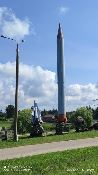 Two missiles and other military equipment are displayed outdoors under a clear blue sky with scattered clouds. The larger missile, grey with an orange base and red tip, stands upright. To its left, a smaller missile, white and blue with fins, is mounted on a wheeled platform. In the background, trees and buildings are visible. A lamppost stands to the left of the frame. The image appears to be taken during the daytime, with bright sunlight. The metadata indicates the image was taken on July 15, 2026, at 10:29 AM by a REDMI NOTE 9 PRO AI QUAD CAMERA.