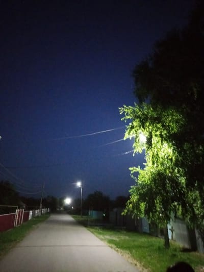 The image captures a nighttime street scene in Voroshilovka, Kazakhstan. A paved road runs through the center of the frame, illuminated by streetlights. To the right, a large, leafy tree is brightly lit by a nearby streetlight, its leaves glowing green. The sky above is a deep, dark blue, with faint white streaks that appear to be contrails from an aircraft. On the left side of the road, a red fence runs along the edge of a property, with a few indistinct structures behind it. Further down the road, more streetlights are visible, casting pools of light on the pavement. The overall impression is one of a quiet, suburban street at night.