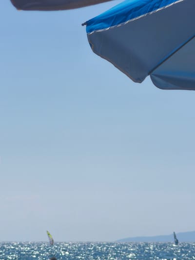 The image shows a view of the sea from a beach in Castiglione della Pescaia, Italy. A blue and white beach umbrella occupies the top portion of the frame, casting a shadow. The sea is visible in the lower part of the image, with sunlight glinting off the surface. Two windsurfers are in the distance on the water. The sky is clear and blue.
