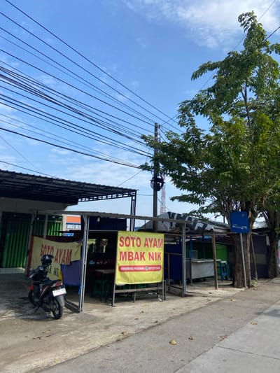 The scene is a street in Kecamatan Tulangan, Indonesia, located at latitude -7.487, longitude 112.654. A large tree with green leaves stands on the right side of the image, casting shade on a food stall to the left. The stall is covered by a yellow awning and has a sign that reads "Soto Ayam Mbak Nik". The stall is empty, with a couple of chairs and a table on the left. A motorcycle is parked in front of the stall. In the background there are several other buildings and stalls, all of which are enclosed by walls. The street is paved with concrete and there are some fallen leaves scattered on the ground. The sky is blue and there are some clouds in the distance. Numerous power lines run overhead, connected to a tall utility pole.  There is a tall red and white radio tower in the background. There are no people in the image.  The image is taken from a street level angle.