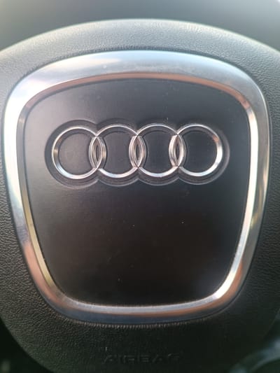 This media file features a close-up, eye-level shot of the Audi logo, which consists of four interlocking rings, embossed on the black airbag cover of a car's steering wheel. The logo is highlighted with a silver-toned trim that encircles it and is further framed by the textured black leather of the steering wheel. The lighting suggests it is daytime, possibly sunny, as indicated by the bright reflections on the metallic trim and the slight glare visible on the dashboard area beyond the steering wheel. The scene is the interior of a car, likely in Germany, given the provided location context of Germersheim. There is no visible activity taking place, and the focus is solely on the car's interior detail. The overall impression is one of manufactured quality and automotive branding. Faint text, possibly "AIRBAG," is visible on the lower portion of the steering wheel, partially obscured by shadow and the texture of the material.