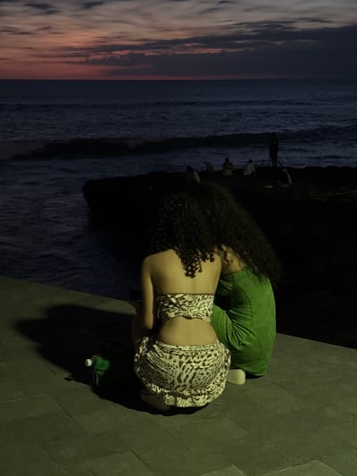 Two women are on a grey concrete path adjacent to a coastline at Pererenan, Indonesia. One woman, in a green garment, is partially visible, lying face down. The other, wearing a light-colored leopard-print dress, is leaning over her, face towards the ocean. The coastline features the ocean on the left and dark cliffs in the center. Several people are on the cliff. There is an orange and grey sunset sky on the left. A green water bottle sits on the path near the women.