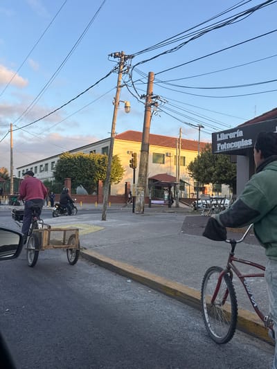 The image shows a street scene in Quilmes Oeste, Argentina, during daylight. A person riding a Vairo-branded bicycle is in the upper right foreground. Next to the bicycle rider is a sign for "Libreria Fotocopias". Further down the street, another person is riding a bicycle that is pulling a cart. A motorcycle is traveling in the same direction. Utility poles with electrical wires dominate the left side of the frame, reaching into the sky. The building in the background has a light-colored facade and a red-tiled roof. The sky is pale blue with some clouds visible. The street is paved, and a yellow curb runs along its edge.