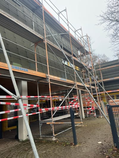 The image displays a multi-story building undergoing construction or renovation, situated in Tuttlingen, Germany. The building's upper levels are clad with dark gray shingle-style siding, featuring visible windows that reflect an overcast sky. Extensive, multi-tiered scaffolding constructed from metal poles and orange wooden planks, some bearing the "BECKER" brand, covers the entire visible facade. The building's ground level incorporates light-colored structural columns and a wooden ceiling, with yellow-framed doorways visible behind safety barriers. Red and white striped barrier tape demarcates an area on the ground in front of the building. The immediate foreground consists of muddy earth with scattered debris and leaves, bordered by a dark gray metal mesh fence with an open gate section on the right. A paved area is visible closer to the building on the left. Bare tree branches are visible against the light gray, overcast sky on the right side of the frame. No individuals are present within the scene.