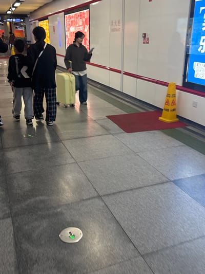 The image shows a busy metro station with passengers walking and pulling luggage. A yellow caution cone stands on a red mat against a white wall with signage. People are waiting or passing through, their attire ranging from casual to formal. A green arrow indicator is embedded in the grey tiled floor. The overall scene suggests a routine day at a transportation hub.