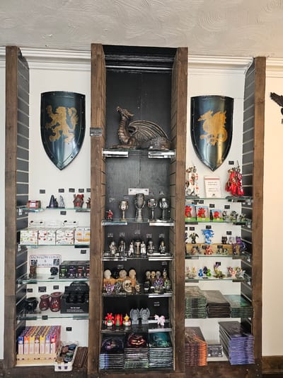 A retail display within a shop in Llandudno Junction, United Kingdom, features three rows of wooden shelves mounted on a wall. The shelves contain an assortment of fantasy and occult-themed items. The top shelf includes toy knights on horseback, small dragon figurines, and small canvas pictures. The middle shelf displays dragon statuettes, goblets, skulls, and fantasy-themed pictures. The bottom shelf contains shields with dragon emblems, incense cones, backflow burners, and manga books. Signs with prices are visible throughout the display.