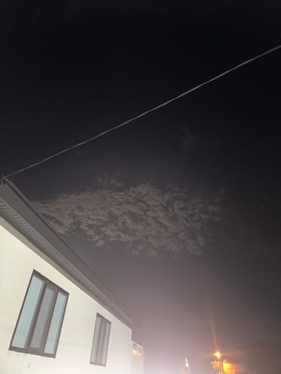 The image captures a nighttime scene in Subutli-Miatli, Russia. A portion of a white building with two windows is visible in the lower left, with a dark roofline extending diagonally across the frame. Above the building, a thin, white electrical wire stretches across the dark sky. Scattered, wispy clouds are illuminated faintly in the mid-sky, suggesting some atmospheric diffusion of light. In the lower right, a bright, orange street light casts a glow, and some indistinct structures are visible behind it. The overall atmosphere is dark and slightly hazy.