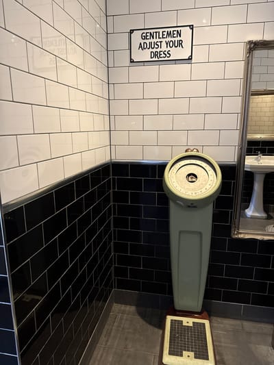 The image depicts a bathroom scene, possibly in a public establishment, with a distinct black and white tile aesthetic. A vintage-style mechanical scale, painted a light green, is the central object. Its dial is visible with numerical markings, suggesting it's functional. To the left of the scale, a sign with a black border reads "GENTLEMEN ADJUST YOUR DRESS" in white capital letters. Behind the scale, a large, ornate mirror reflects a sink and more tiling, indicating the presence of other bathroom fixtures. The floor is a mix of black and white subway tiles, with a grey tiled area visible to the far right. The lighting suggests an indoor setting, likely during the day, given the bright reflection in the mirror. There are no people visible in the image. The overall impression is one of a retro-themed or historical bathroom. The location context provided, Poole, United Kingdom, adds a specific geographical reference to this domestic or public space.