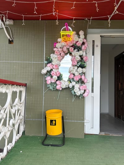 The image depicts an exterior view of a building with several decorative elements. A floral wreath, composed of white and pink roses, hangs above a white railing. To the left, a neon sign depicting a bubble tea drink is visible, and a string of lights adorns a red wall. To the right, there is a yellow trash receptacle with a graphic symbol on its side. The setting appears to be urban, but there is no definitive evidence to identify the location specifically as Uray, Russia.