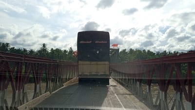 The image shows the rear of a covered truck driving on a bridge. The truck has two flags on it, both red and white. The bridge is made of red metal beams and has a rough asphalt surface. Palm trees and other vegetation can be seen on the banks of the river below the bridge. The sky is cloudy. The location is Awegeutahpaya, Indonesia.