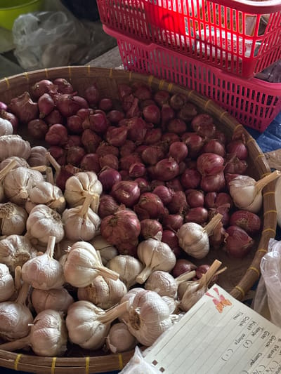 A close-up, overhead shot captures a woven basket filled with produce, predominantly red onions and heads of garlic. The basket is positioned on a blue surface, possibly a market stall, with two stacked, bright red plastic baskets visible in the upper right corner. The lighting is slightly dim, highlighting the textures of the vegetables. In the foreground, a corner of a notepad with handwritten Indonesian text and a small sticker of an orange butterfly is visible, suggesting this scene is within Karangsambung, Indonesia, a location known for its agricultural produce markets. The overall impression is a market scene focused on fresh ingredients.