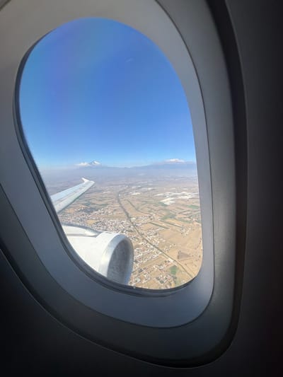 The image displays an aerial view captured from an aircraft window, showcasing the landscape in and around Santa Ana Xalmimilulco, Mexico, under a clear, bright blue sky. The dark grey, contoured frame of the airplane window occupies the left and bottom portions of the foreground. A silver-grey aircraft wing extends into the frame from the left, and a section of a matching engine nacelle, with its circular intake visible, is situated below the wing in the lower-right. The ground below consists of a varied terrain featuring a patchwork of agricultural fields, ranging from brown to green, interspersed with numerous clusters of residential and commercial structures indicative of urban and suburban development. Linear features, possibly roads or canals, are discernible crisscrossing the landscape. In the distant background, two prominent snow-capped mountain peaks, consistent with Popocatépetl and Iztaccíhuatl, are visible rising above a noticeable layer of atmospheric haze. No human figures or explicit actions are depicted.