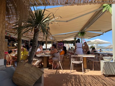 This is a medium shot of a beachside restaurant in San Roque, Spain, on a sunny day. The scene is outdoors, with a thatched roof and a large beige awning providing shade over several tables. The ground is covered in reddish-orange sand. Several people are seated at tables, dining and socializing. In the foreground, a woven straw bag sits on a low, grey upholstered bench. Palm trees and ferns add to the tropical ambiance. In the background, beach umbrellas and the sea are visible. The atmosphere appears relaxed and convivial, typical of a coastal dining establishment.