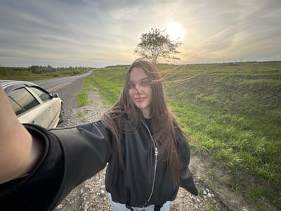 A woman stands near a car on a road in a rural area of Бикбарда, Russia, during the daytime. She faces the camera wearing a black leather jacket and jeans. The location appears to be a roadside with grass fields and a solitary tree between the road and sky. The sky is visible, with some cloud cover and bright sunlight.