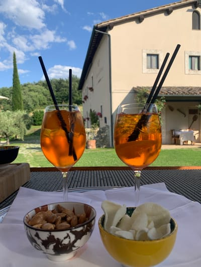 The image depicts two Aperol Spritz cocktails served in wine glasses with black straws and ice, placed on a table with a grey placemat. In the foreground, two small bowls are visible: one contains almonds, and the other contains white crisps. In the background, a building with a tiled roof and pale yellow walls stands against a backdrop of lush greenery, cypress trees, and a bright blue sky with scattered clouds. The setting suggests an outdoor dining area, possibly at a restaurant or villa in the Tuscan countryside. The overall scene conveys a relaxed and pleasant atmosphere.