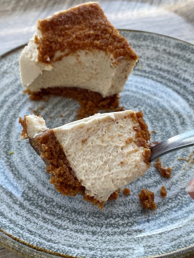 This media file captures a close-up view of two slices of cheesecake on a speckled gray plate. A fork is in the process of being used to lift a portion of one of the slices. The cheesecake has a creamy white filling and a brown, crumbly crust. The plate itself has a textured, speckled pattern and a slightly raised brown rim. The background is softly blurred, suggesting natural light, possibly from a window. The overall scene is casual and focuses on the dessert, hinting at a meal or a snack. The presence of a hand and fork indicates someone is about to eat or is in the process of eating the cheesecake. The lighting suggests it might be daytime. Given the location context of Algeciras, Spain, it's possible this is a dessert enjoyed at a restaurant, cafe, or home in that area. There is no visible text in the image.