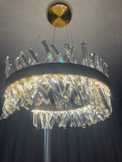 A circular chandelier, adorned with numerous clear crystal prisms, hangs from a gold-colored ceiling mount in the city of Moni Community, Cyprus. The prisms are arranged in a radiating pattern, creating a sparkling effect with visible rainbow refractions from the embedded lights. The chandelier is suspended by thin wires against a dark, draped background.
