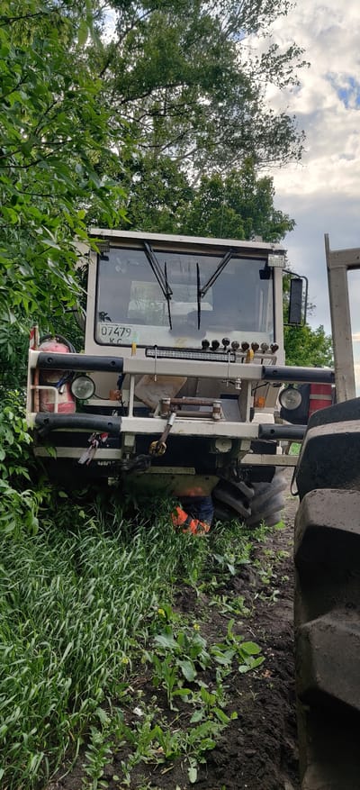 The image shows a white tractor partially off the road in Kovalivs'ke, Ukraine. The tractor's front tires are in the dirt and grass, and a large tire is visible on the right side. The tractor has a cabin with windshield wipers and a license plate with "07479 KC" on it. The tractor is situated amidst a field of green grass and foliage with soil visible. There are trees and a cloudy sky in the background. A person wearing orange can be seen working under the tractor.