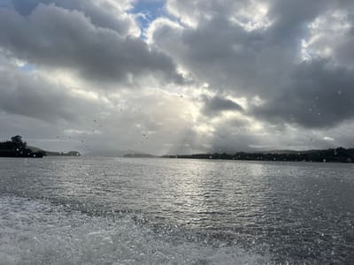 The image presents a wide view of the Minho River estuary in the vicinity of Vila Nova de Cerveira, Portugal. The sky is predominantly overcast with heavy, dark grey clouds, yet shafts of sunlight penetrate through a central break, illuminating a portion of the water. The river's surface is dark, rippled, and reflects the subdued light from above. In the immediate lower left foreground, a significant white wake of disturbed, frothing water is visible, indicating the movement of a vessel from which the photograph was taken. Numerous small, translucent water droplets are suspended across the mid-ground, suggesting precipitation or spray. The distant horizon is defined by a continuous, dark line of densely forested hills. A closer landmass featuring dark green vegetation and a subtle, dark, possibly structural, element is discernible on the left mid-ground. No individuals, identifiable vessels other than the implied point of view, or specific interactions are visible.