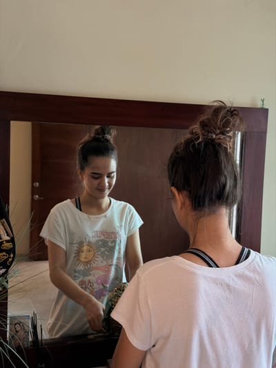 A woman with brown hair in a bun leans over a dark wood-framed mirror. Her reflection shows her wearing a white t-shirt with a sun graphic and the words "JUST LET GO." Her back is to the viewer, she is wearing a white t-shirt with a black and white striped bra. The room has pale walls. A bag and plants are near the bottom of the mirror.