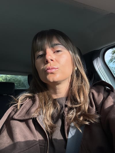 A young woman is sitting in a car. She has brown hair with blonde highlights and brown eyes, wearing a brown jacket and a seatbelt. The car window shows green vegetation and sunlight, suggesting an outdoor location. The scene appears to be taken during daylight. This cannot be identified as being in Mandelieu-la-Napoule, France from the given image and details.