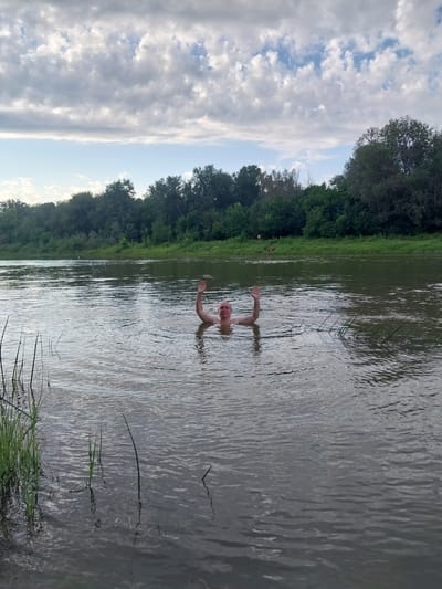 A bald man is swimming in a body of water, with his arms raised and hands waving. He is partially submerged, with his head and shoulders visible above the water. The water is calm and appears to be a river or lake. In the background, there is a dense line of trees and vegetation along the shoreline. The sky is cloudy with some patches of blue visible. The scene is outdoors, likely during the day. The location is the bank of the Great Chkalov River in the city of Chkalov, Russia.