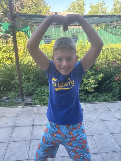 A young boy with blond hair and a wide smile is posing outdoors, forming a heart shape with his arms above his head. He is wearing a blue t-shirt with a yellow logo that appears to be "Carlsberg" and blue shorts with an orange cartoon character print. The background shows a lush garden with green foliage and some pink flowers. The ground is paved with gray square tiles. A wooden pole and a green mesh structure are also visible in the background. The scene suggests a warm, sunny day in a residential outdoor setting, likely in Zmiiv, Ukraine, given the context.