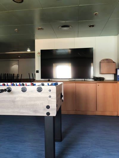 The image shows an interior setting. On the wall is a flat screen television mounted between a light olive green wall with ceiling-mounted lights and a wooden cabinet. A foosball table sits in front of the television. The floor is a deep blue color. The scene appears to be indoors. No people are visible. It's impossible to confirm the location of La Spezia, Italy, without additional context.