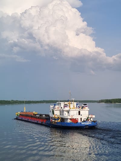 A barge navigates a body of water. The barge is carrying a load of wrapped materials. The tugboat accompanying the barge is white with a blue hull. The scene takes place on a river in a location that appears to be Shelomovo, Russia. The water is calm, and there are trees lining the banks in the distance. Above, there is a cloudy sky.