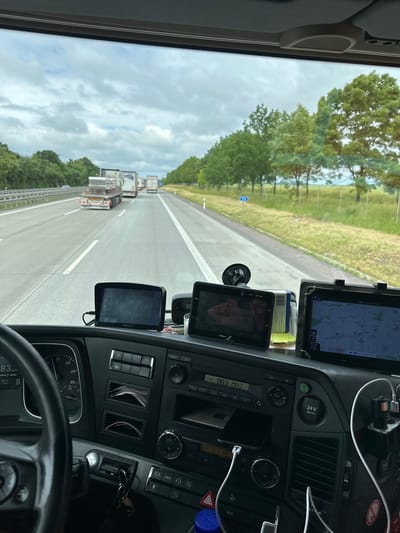 The image is taken from the driver's perspective inside a truck cab, looking forward through the windshield. The truck is on a highway, with other trucks in front of it. The weather is overcast. The dashboard is visible, featuring a steering wheel, various buttons and controls, and two GPS devices mounted on the windshield. One GPS shows a map with the route, indicating a location near "Erfurt Chemnitz". The other GPS has a brand name "GBox". The dashboard also displays a speedometer reading 83 km/h. The environment outside the truck consists of a highway, trees and vegetation on the sides, and a cloudy sky. The scene suggests the truck is in motion on a road in Germany.