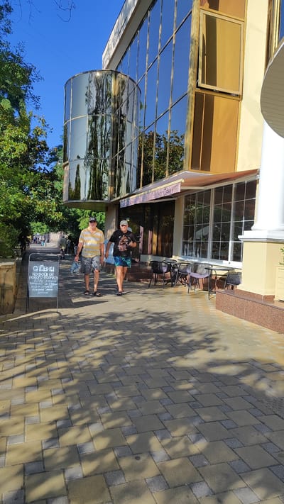 Two men walk down a sidewalk in front of a modern building with reflective gold-tinted windows and a cylindrical glass facade. The man on the left wears a yellow and white striped polo shirt and grey shorts, carrying a plastic bag. The man on the right wears a black t-shirt with a graphic print and multi-colored shorts, holding a black object in his right hand. A chalk A-frame sign in front of them displays Russian text. The sidewalk is paved with interlocking yellow and grey pavers, and shadows from trees are cast across it. The building appears to be a commercial establishment, possibly a cafe, with outdoor seating visible through its large windows. The environment suggests a sunny day in a town or city, possibly along a promenade or street.