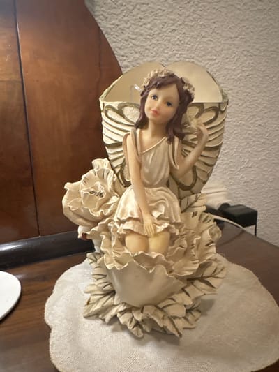 The image shows a close-up, eye-level shot of a decorative figurine of a young girl with wings, resembling an angel or fairy. She is seated within a large, stylized flower with multiple layered petals in shades of cream and beige. The figurine has brown hair adorned with a floral wreath, blue eyes, and is wearing a light-colored, ruffled dress. Her right hand is raised, holding a small, light-colored object, possibly a bird or a flower. The figurine is placed on a round, light-colored placemat with a scalloped edge, which sits on a wooden surface. Behind the figurine, a decorative lamp shade or backing with ribbed details in cream and gold is visible. To the left of the figurine, a portion of a dark brown wooden piece of furniture, possibly a cabinet or desk, is present. To the right, a textured white wall and a black electrical outlet with a cord are discernible. The lighting is soft, casting some shadows. The scene is indoors, and the overall impression is of a decorative tabletop item. No specific location in Santa Pola, Spain, can be identified from the image.