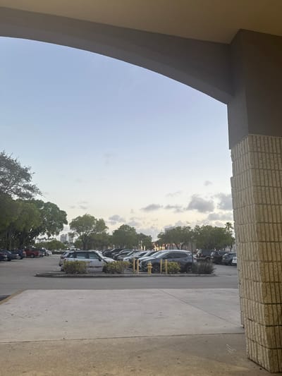 The image depicts an outdoor parking lot scene in Aventura, United States, viewed from beneath a grey-painted concrete archway and beside a light-colored, fluted stone block pillar on the right. The sky is clear light blue with scattered white and grey clouds, indicating daylight, likely late afternoon or early evening.
The foreground consists of a flat, light-grey concrete surface. The middle ground and background are occupied by a large asphalt parking lot containing numerous parked vehicles arranged in rows. A light-colored SUV, possibly silver or light grey, with a horizontal red marking or reflection on its driver's side, is prominently parked near a landscaped island in the center-left. This island features low green bushes, a yellow fire hydrant, and several yellow bollards. Other vehicles visible include multiple dark-colored sedans and SUVs, and a red pickup truck in the far-left background.
The parking lot is bordered and interspersed with mature green trees, including tall palm trees and various leafy trees, particularly on the left and along the distant perimeter. In the far background, beyond the trees, several multi-story buildings and urban structures are discernible. No people are present or engaged in any activities within the visible frame.