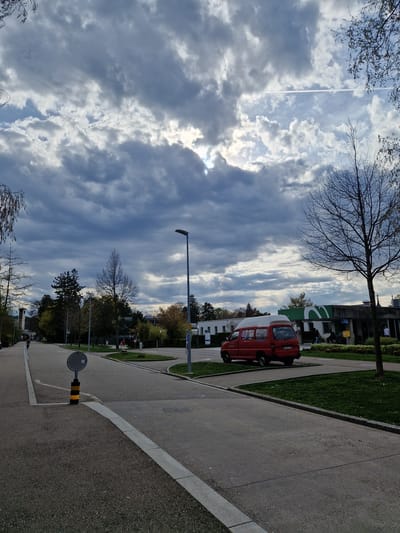 The image shows a street scene in Rheinfelden, Switzerland. A red and white compact utility van is parked on the side of a paved road with adjacent grass and sidewalk. The sky is partially cloudy with contrails visible. There is a street lamp and a convex mirror on a pole near the road. Trees with bare branches are visible, along with other trees and buildings. No people are readily visible.