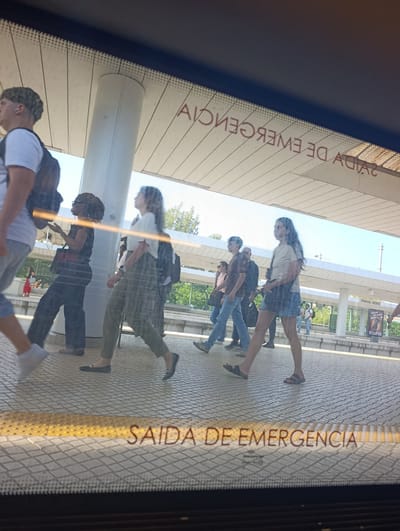 In Pinhal Novo, Portugal, a platform at a train station is visible through a reflective surface, likely a window. Several people are walking on the platform. One person in the foreground on the left is wearing a white t-shirt and dark pants. To their right, a person in dark clothing walks with a red bag over their shoulder. Another person in a light-colored t-shirt and green pants is walking forward. Further down the platform, a group of people are walking. One individual is wearing a brown t-shirt and blue jeans, accompanied by others. In the background, a sign reads "PINHAL NOVO" and below it an advertisement for "ECLIPSE". The word "SAIDA DE EMERGENCIA" (Emergency Exit in Portuguese) is clearly visible on the textured yellow and grey platform surface in the foreground. The scene appears to be captured during daylight hours.
