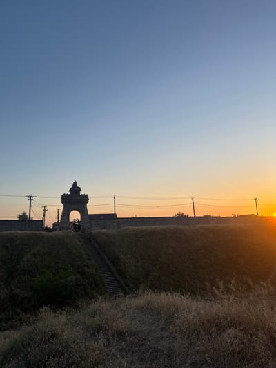 The image captures a sunset in Mizhlymanske, Ukraine, featuring a stone archway and a long set of stairs leading up a grassy hill. The sky transitions from a clear blue at the top to a warm orange glow near the horizon, where the sun is setting. A concrete wall runs along the top of the hill, with power lines and poles visible against the sky. Several people are seen walking up the stairs towards the archway. The foreground is dominated by dry, golden grass.