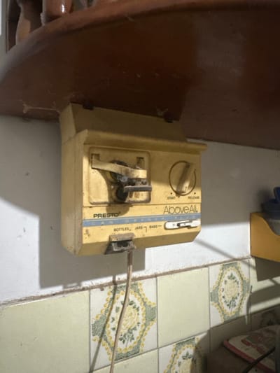 A yellow, aged Presto Above All can opener sits on a white surface adjacent to a wooden cabinet. The device, marked with "START," "RELEASE," "BOTTLES," "JARS," and "BAGS," has a dull appearance. A power cord extends from its side. The surface the can opener is on appears to be tiled, with a yellow and green floral pattern visible in some of the tiles. The image appears to be taken in a cluttered environment, suggesting the can opener has been in use for a long time.