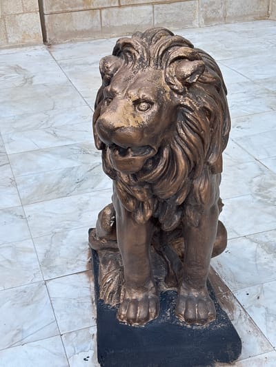 A bronze-colored lion statue is positioned on a dark rectangular base, which rests on a tiled floor. The statue is seated with its front paws forward and its tail curled around its side. The lion has a detailed mane, textured fur, and an open mouth. The surrounding floor is tiled with a white marble pattern, and behind the statue, a wall constructed of light-colored rectangular blocks is visible. The overall scene appears to be outdoors in a public space in Al Qastal, Jordan.
