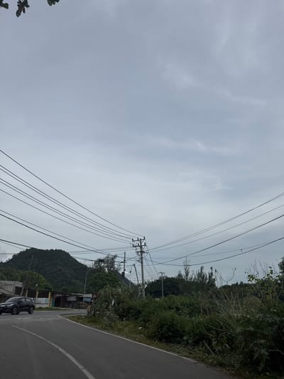 The image shows a view of a street with a car and vegetation in Lamkruet, Indonesia. A black car is driving on the left side of a paved road. The road has white lane markings. On the left side of the road, there are some low-lying structures and a prominent green hill in the background. Overhead, multiple electrical wires are suspended from utility poles. The sky is overcast and gray. The vegetation consists of various trees and bushes, some of which are tall and dense, while others are smaller and more sparse. In the distance, more trees and hills are visible. The overall scene suggests a somewhat rural or semi-urban environment.