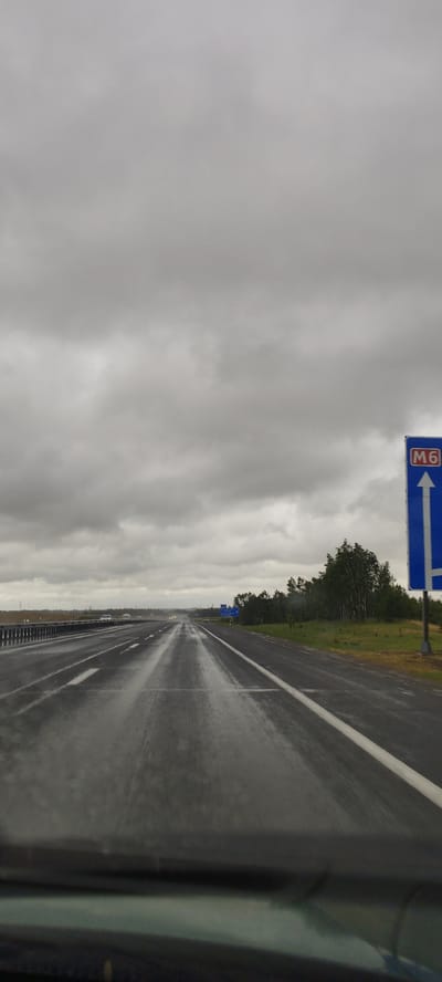 The image shows a view from inside a vehicle driving on a wet highway. The road is glistening with water, indicating recent rain. The sky is heavily overcast with dark grey clouds, contributing to the dim lighting conditions. In the distance, the highway stretches out and appears to curve slightly to the left. There are white lane markings visible on the road. On the right side of the highway, there is a blue directional sign with a white arrow pointing upwards, indicating "M6". Beyond the road and the sign, there is a line of trees and some grassy areas. Several cars are visible on the highway in the distance. The lower portion of the image shows the dashboard and windshield of the vehicle from which the photo was taken. The location is identified as the M6 highway near Путрышкаўскі сельскі Савет, Belarus.