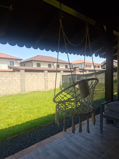 The image depicts a tranquil outdoor scene from a covered porch area, looking out onto a backyard in Dumbravita, Romania. A macrame hammock chair, adorned with a light-colored towel, hangs from the porch ceiling by thick ropes. The porch floor is tiled with dark gray, wood-effect tiles, and edged with a reddish-brown stone border. Beyond the porch, a lush green lawn stretches towards a concrete block wall. The lawn is bordered by a strip of dark gray decorative gravel. In the background, two modern houses with terracotta tile roofs are visible, separated by the concrete wall. The sky is clear and blue, suggesting a sunny day. The overall atmosphere is one of peaceful relaxation and domestic comfort.