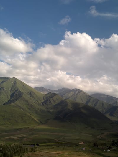 This media file captures a dramatic mountain landscape, likely in Karagai Bulak, Kyrgyzstan. The scene is divided by a clear diagonal line, with the left side dominated by a vibrant blue sky filled with fluffy white clouds. The right side features imposing, verdant mountains that rise steeply. These mountains are covered in lush green vegetation, with visible terraced fields and a small cluster of houses suggesting human habitation and agricultural activity. The terrain appears rugged and uneven, with deep ravines and sharp ridges.
The sunlight suggests it is daytime, likely late morning or early afternoon, given the brightness of the sky and the shadows cast on the mountains. The weather appears fair, with a mix of sun and clouds. The overall mood is one of natural grandeur and perhaps a sense of isolation due to the remote mountainous setting. No people are visible, nor are there any distinct objects that draw attention apart from the natural elements and the distant dwellings. There is no visible text in the image.