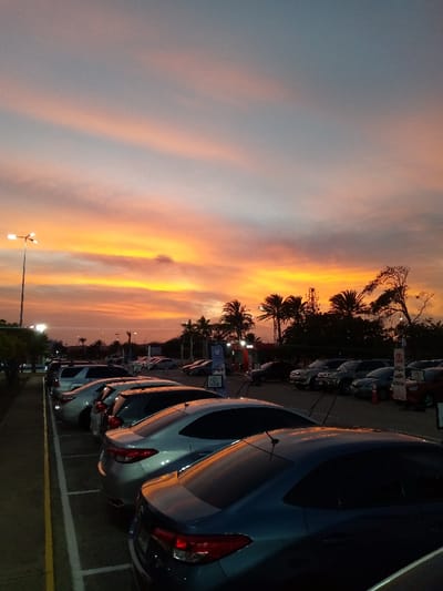 The image displays a stationary outdoor scene within a parking lot at sunset, located in La Cruz del Pastel, Venezuela. The sky dominates the upper portion, exhibiting a pronounced color gradient from intense orange and yellow hues near the horizon, transitioning upwards through pink and pale orange, to light blue at the zenith. Diffuse, streaky cloud formations are present across the sky.

A tall streetlamp with multiple illuminated fixtures stands on the far left, providing artificial light. The lower two-thirds of the image show a paved parking area with white painted lines delineating individual parking spaces. Numerous passenger vehicles, primarily sedans and SUVs, are parked in multiple rows, angled slightly away from the viewer. Notable vehicles in the foreground include a dark blue/teal sedan and a silver sedan. All visible vehicles are unoccupied.

In the mid-ground and background, especially on the right side, a dense line of tropical vegetation is visible, characterized by numerous palm trees and other deciduous trees silhouetted against the sunset. Beyond these trees and vehicles, faint outlines of distant structures, consistent with commercial buildings or a gas station, are discernible, accompanied by scattered artificial lights. No individuals, direct actions, or interactions are depicted within the frame.